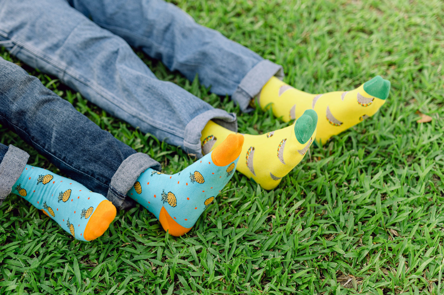One adult wearing pineapple printed socks and another wearing taco printed socks both laying on the grass